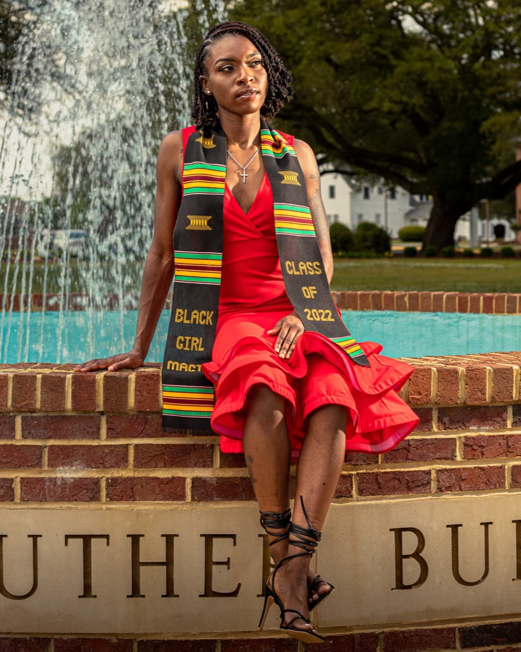 Graduation portrait in a red dress and kente stole by a campus fountain